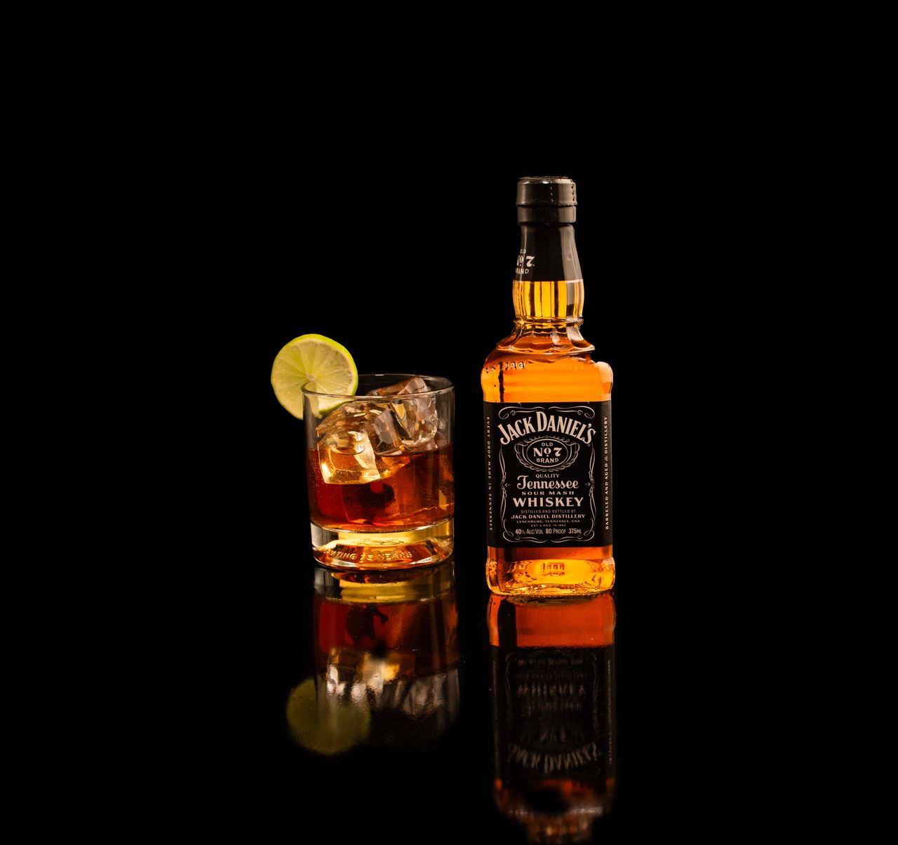 Jack Daniel's bottle and whiskey glass with lime on reflective black surface.